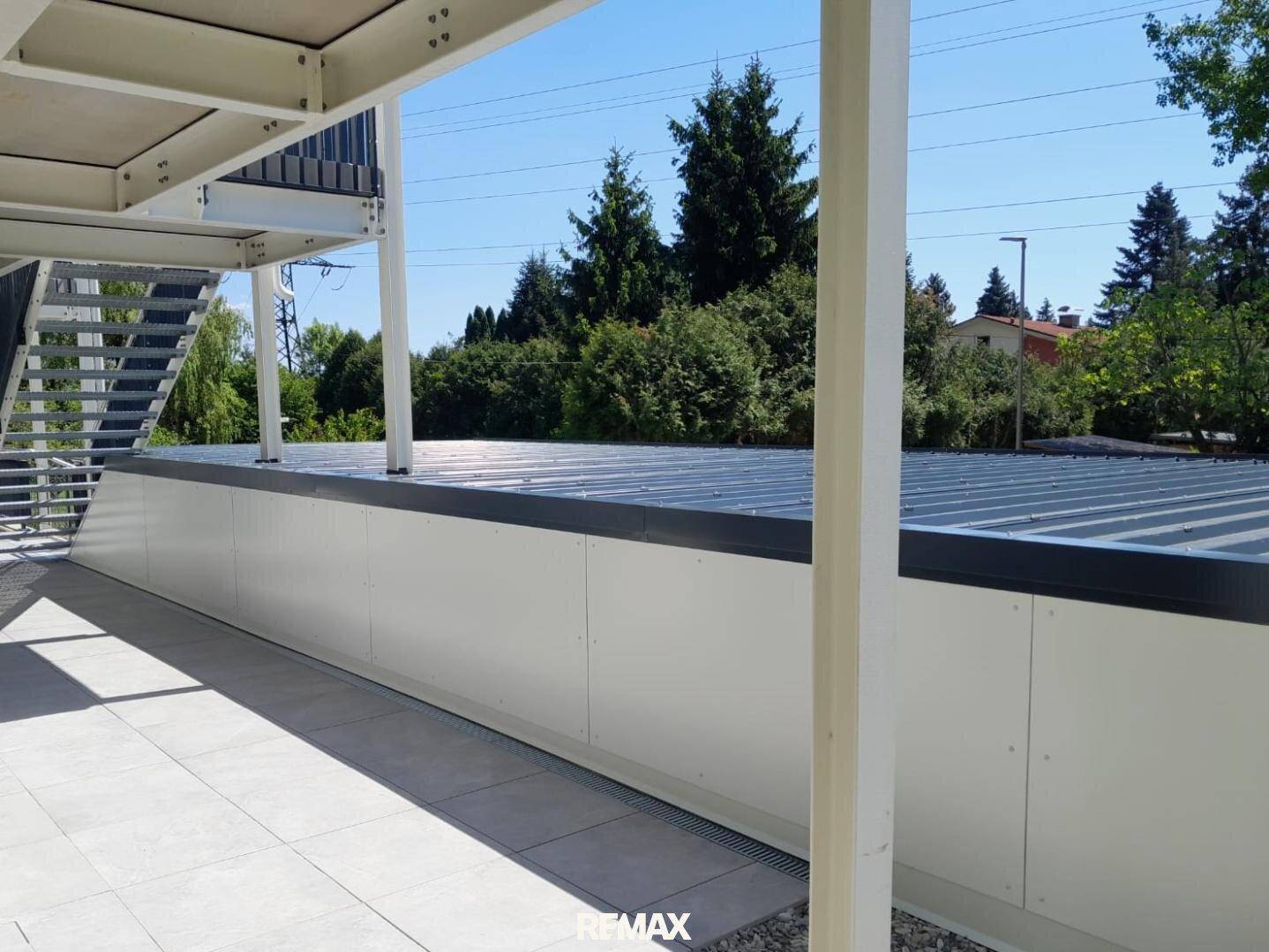 Blick von der Terrasse auf den Carport und die grüne Umgebung unter blauem Himmel.