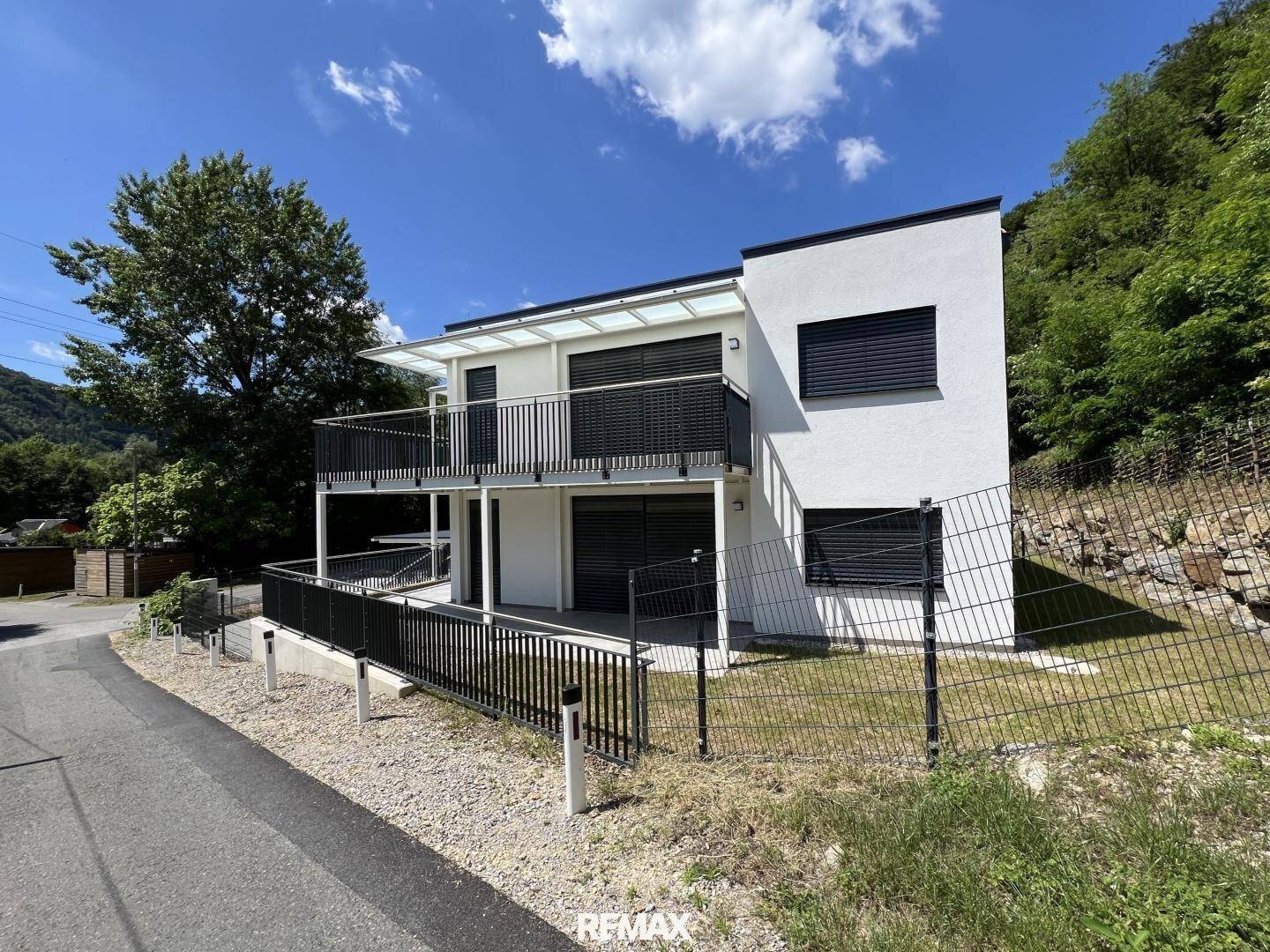Modernes weißes Gebäude mit schwarzen Balkongeländern und Fensterläden, eingebettet in grüne Hügellandschaft.