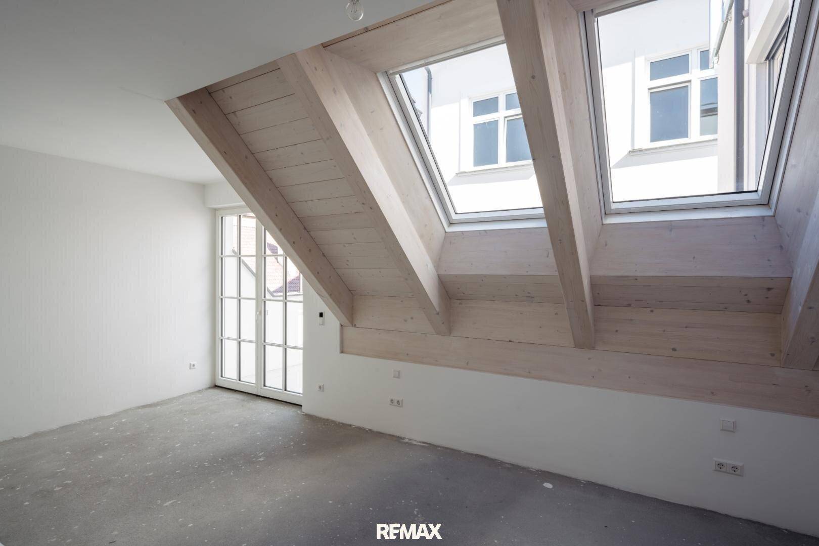 Helles Dachzimmer mit Holzbalken, großen Dachfenstern und Zugang zu einem Außenbereich.