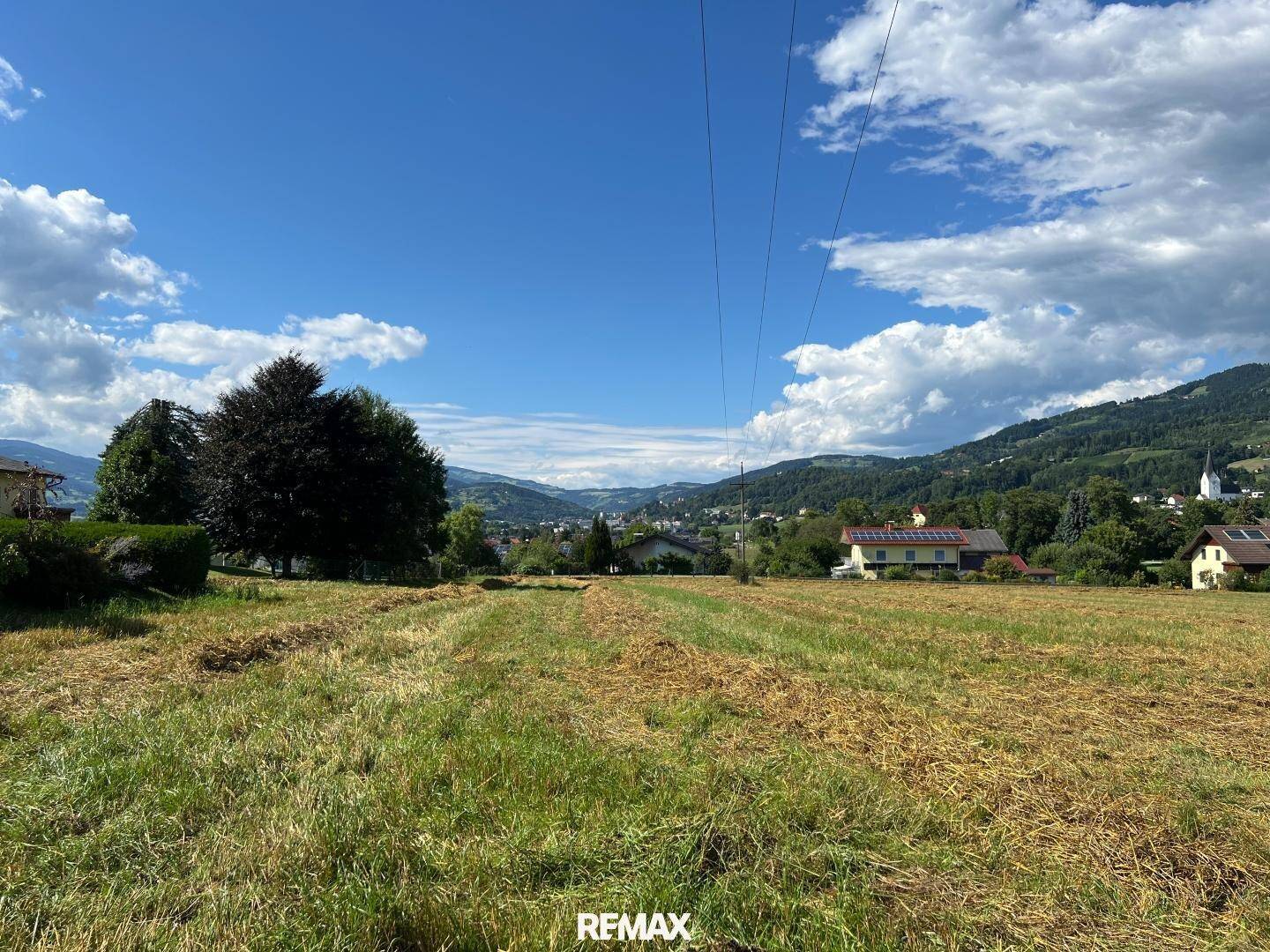 Weitläufiges Feld mit Bäumen, Häusern und Bergen in der Ferne unter einem weiten Himmel.