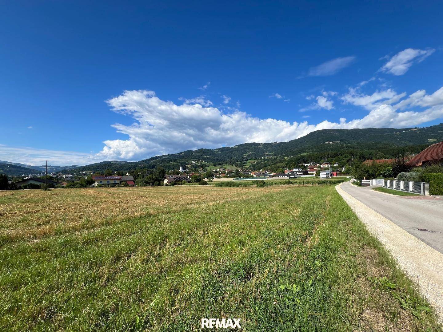 Blick über ein Feld und eine Straße mit Häusern und Bergen im Hintergrund unter blauem Himmel.