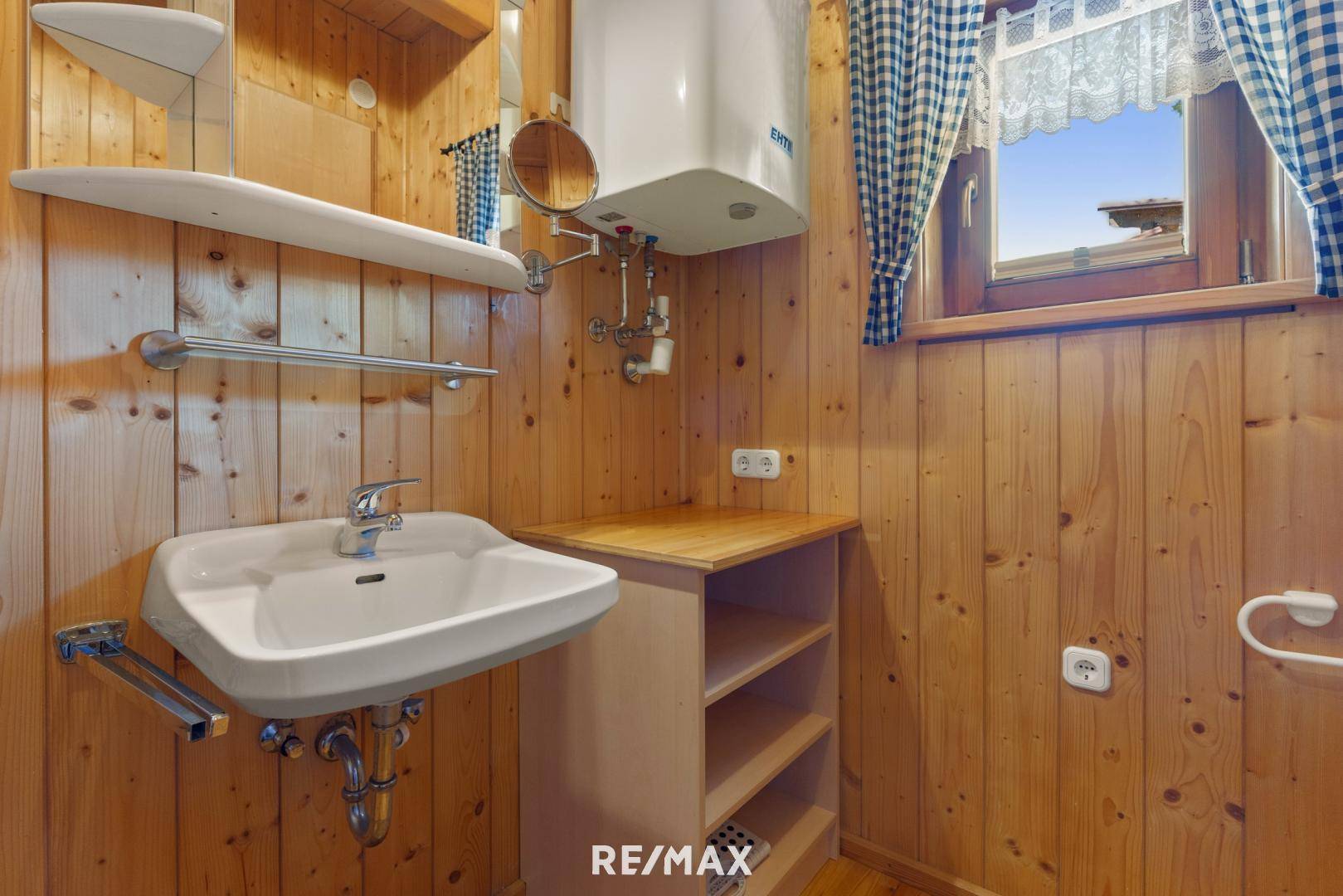 Rustikales Badezimmer mit Holzverkleidung, Waschbecken und Fenster mit karierten Vorhängen, hell beleuchtet.