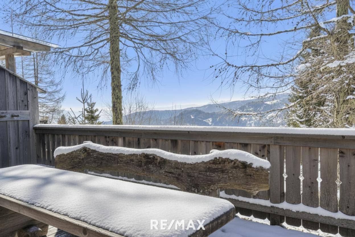 Beschneite Holzbank auf der Terrasse mit weitem Blick über die verschneite Berglandschaft unter blauem Himmel.