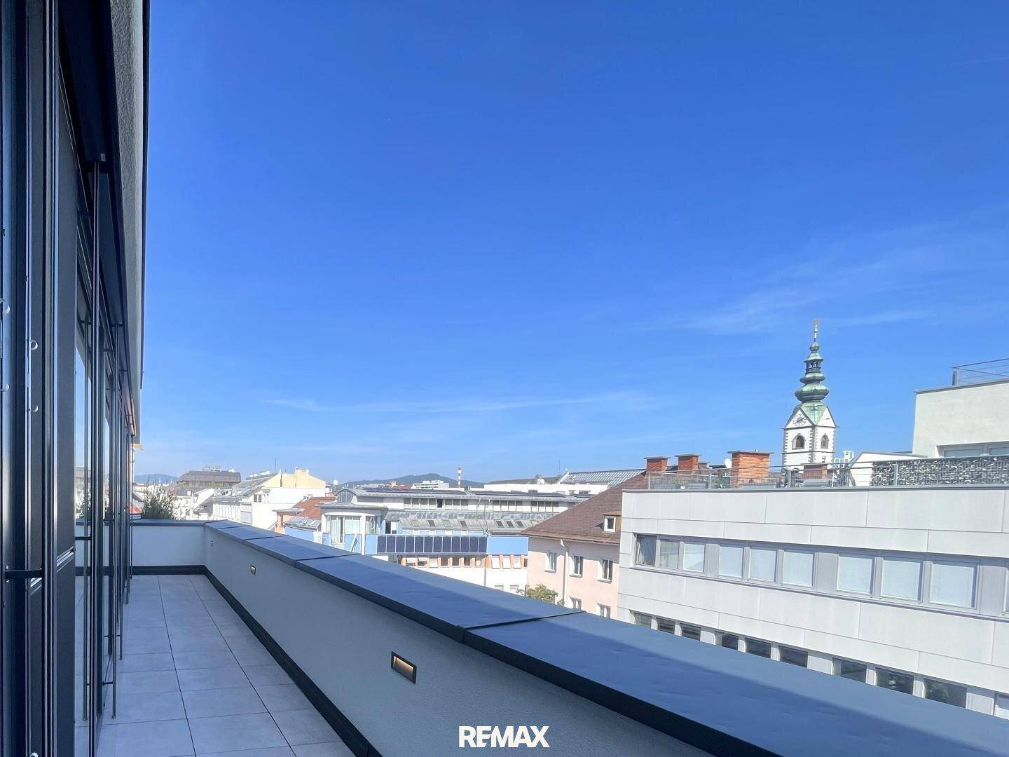 Weitläufige Dachterrasse mit Panoramablick über die Stadt und den historischen Kirchturm unter blauem Himmel.