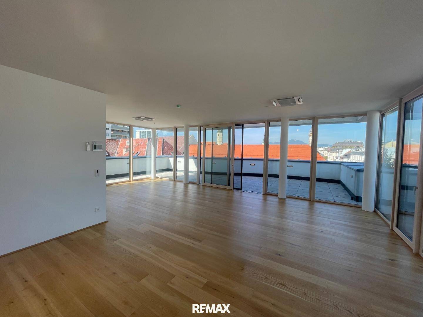 Geräumiger, unmöblierter Wohnbereich mit hellem Holzboden und Panoramafenstern, die auf eine Dachterrasse führen.