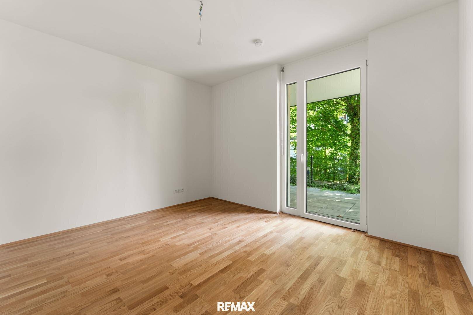 Heller, leerer Raum mit Holzboden und großem Zugang zu grüner Außenfläche.