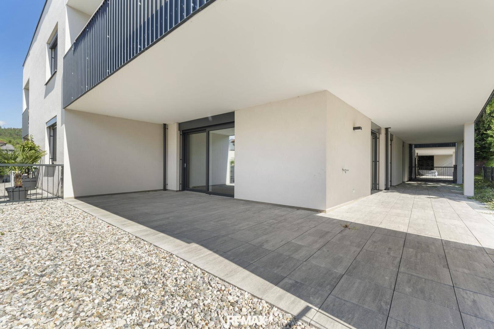 Große, modern gepflasterte Terrasse mit Zugang zum Gebäude und Kiesbereich unter blauem Himmel.
