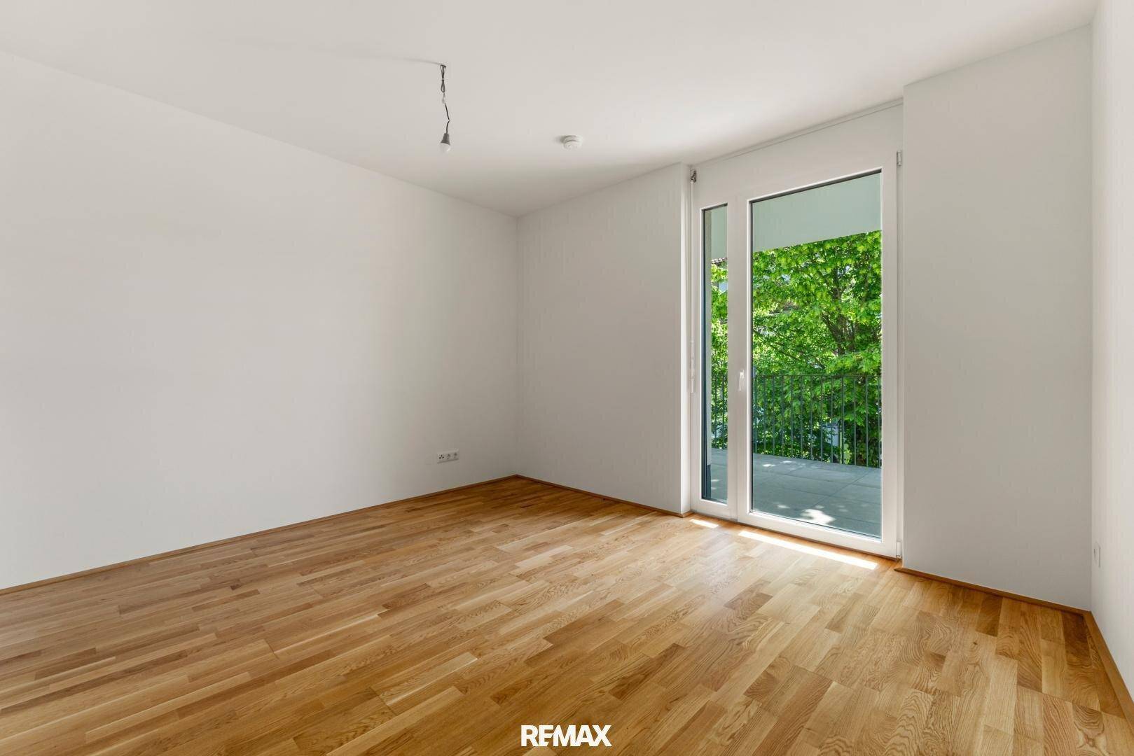 Heller, leerer Raum mit Holzboden und großem Fenster mit Zugang zu einem Balkon und Blick ins Grüne.