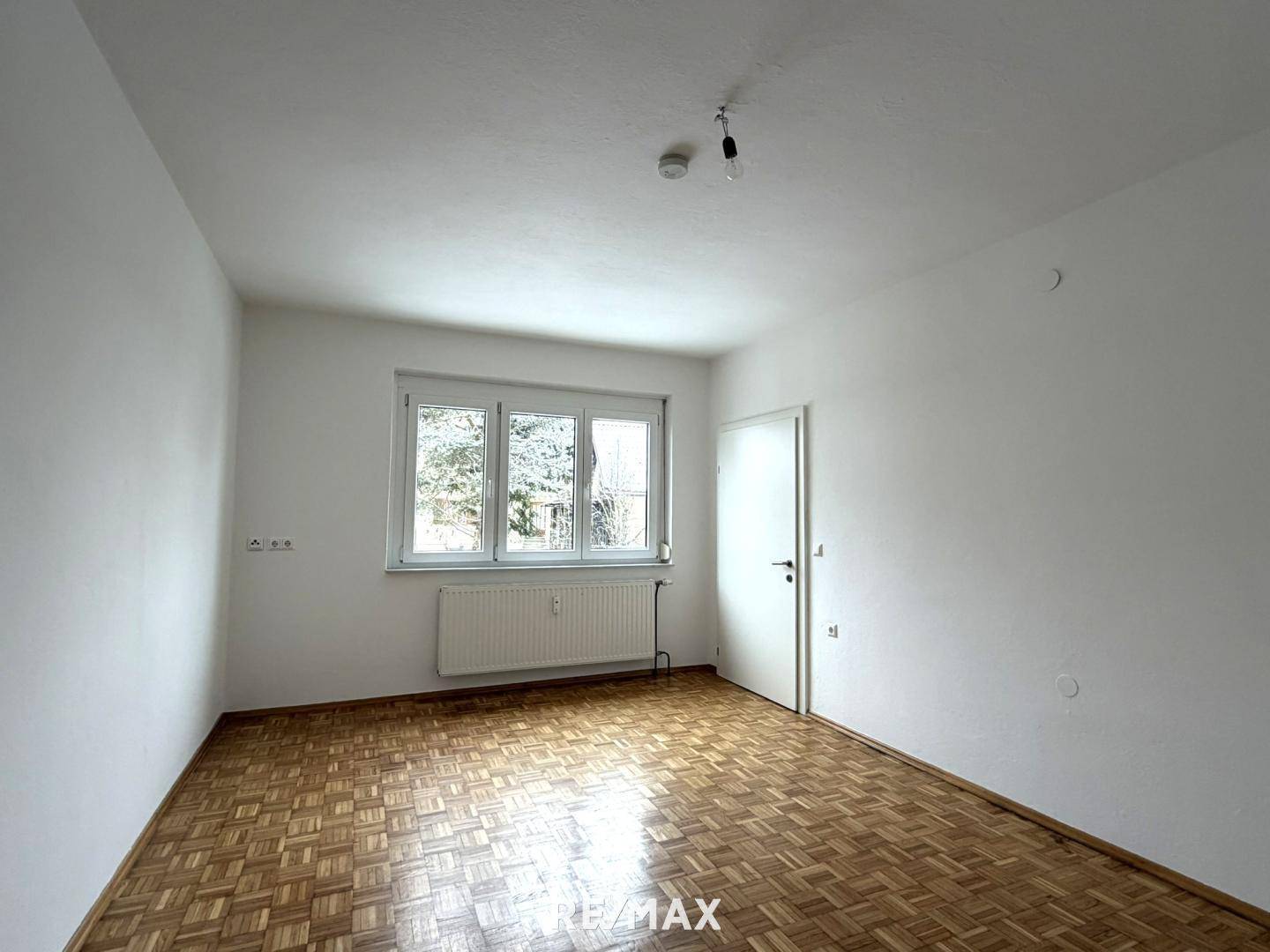 Helles Zimmer mit Parkettboden, großem Fenster und weiß gestrichenen Wänden.