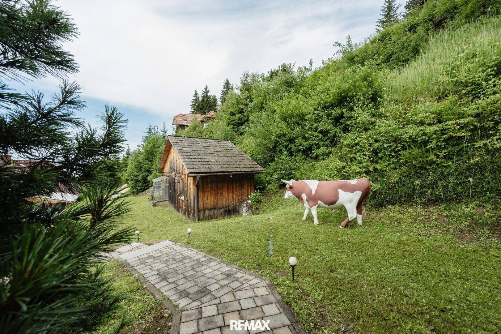 Grüner Gartenbereich mit einem Holzschuppen, einem gepflasterten Weg und einer dekorativen Kuh.