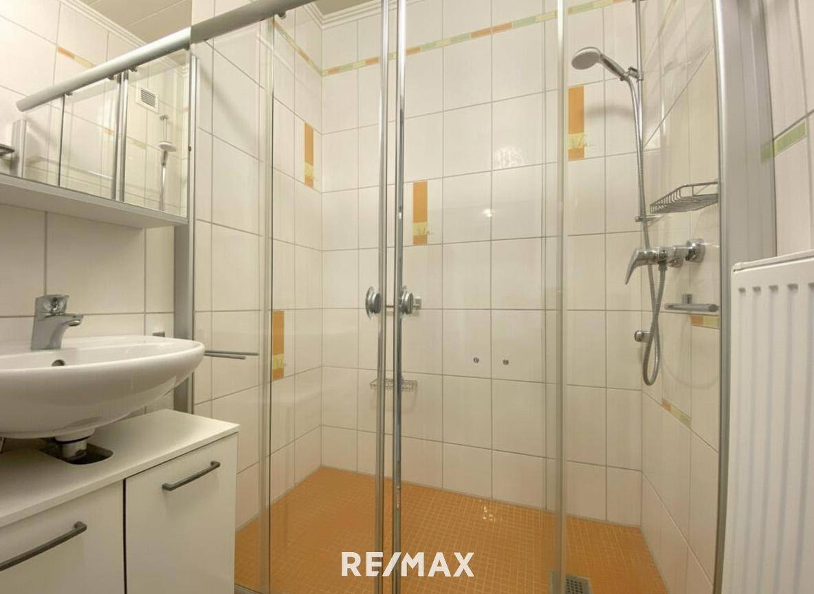 Badezimmer mit Duschkabine, Waschbecken und weißen Fliesen mit orangefarbenen Akzenten.