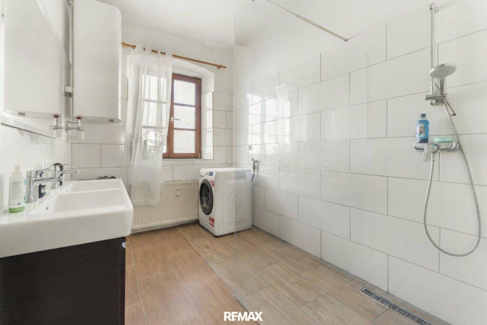 Helles Badezimmer mit weißen Fliesen, zwei Waschbecken und einer ebenerdigen Dusche.