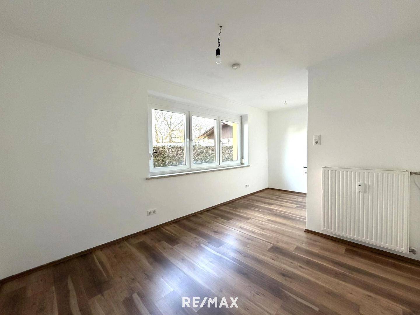 Heller Wohnraum mit Laminatboden und großem Fenster, bietet Blick ins Grüne.