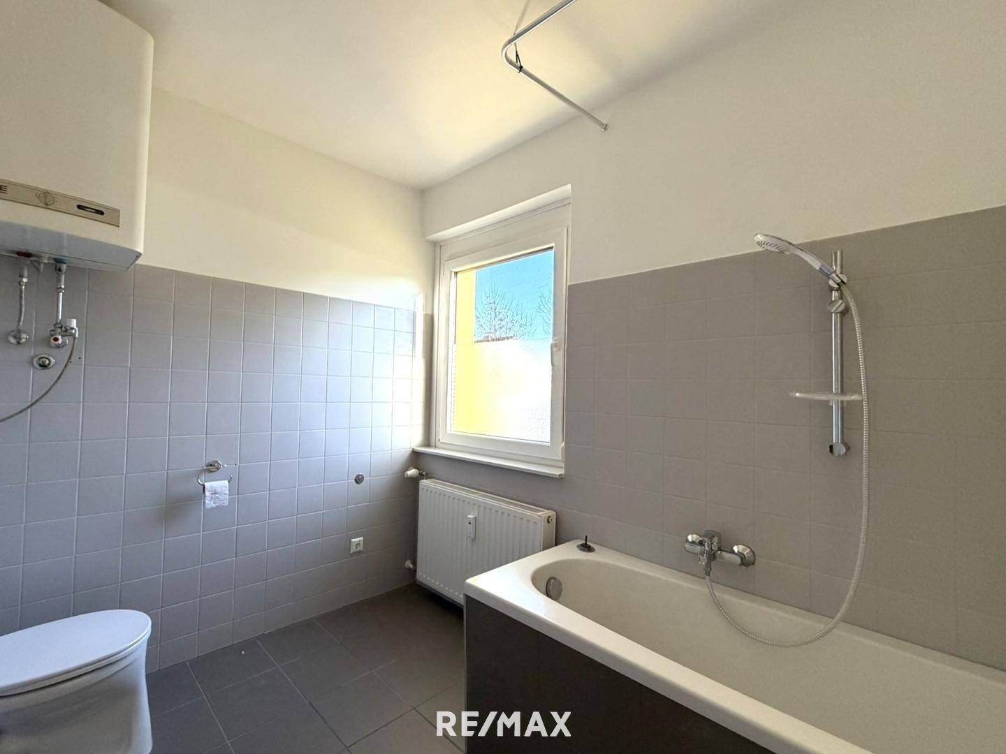 Badezimmer mit Badewanne, WC und Fenster, ausgestattet mit grauen Fliesen.