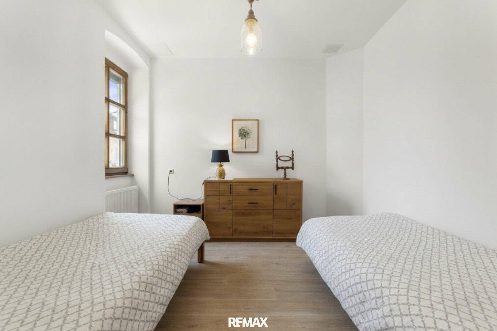 Helles Schlafzimmer mit zwei Einzelbetten, Holzkommode und großem Fenster.