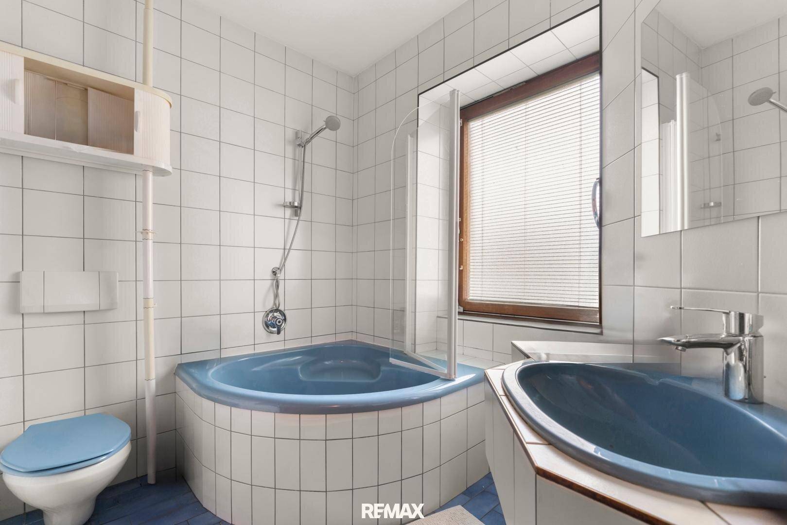 Badezimmer mit blauer Eckbadewanne, Toilette, Waschbecken und Fenster mit Jalousien.