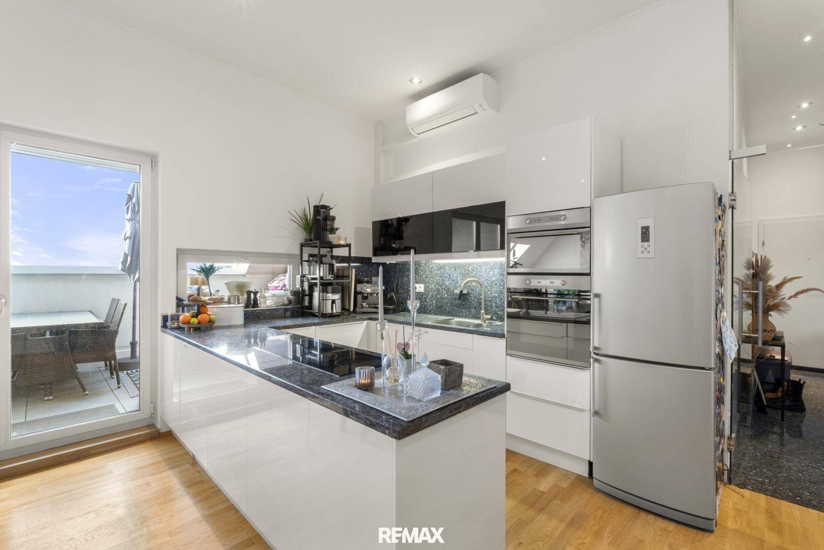 Moderne Küche mit weißen Hochglanzfronten, dunkler Arbeitsplatte und Zugang zum Balkon.