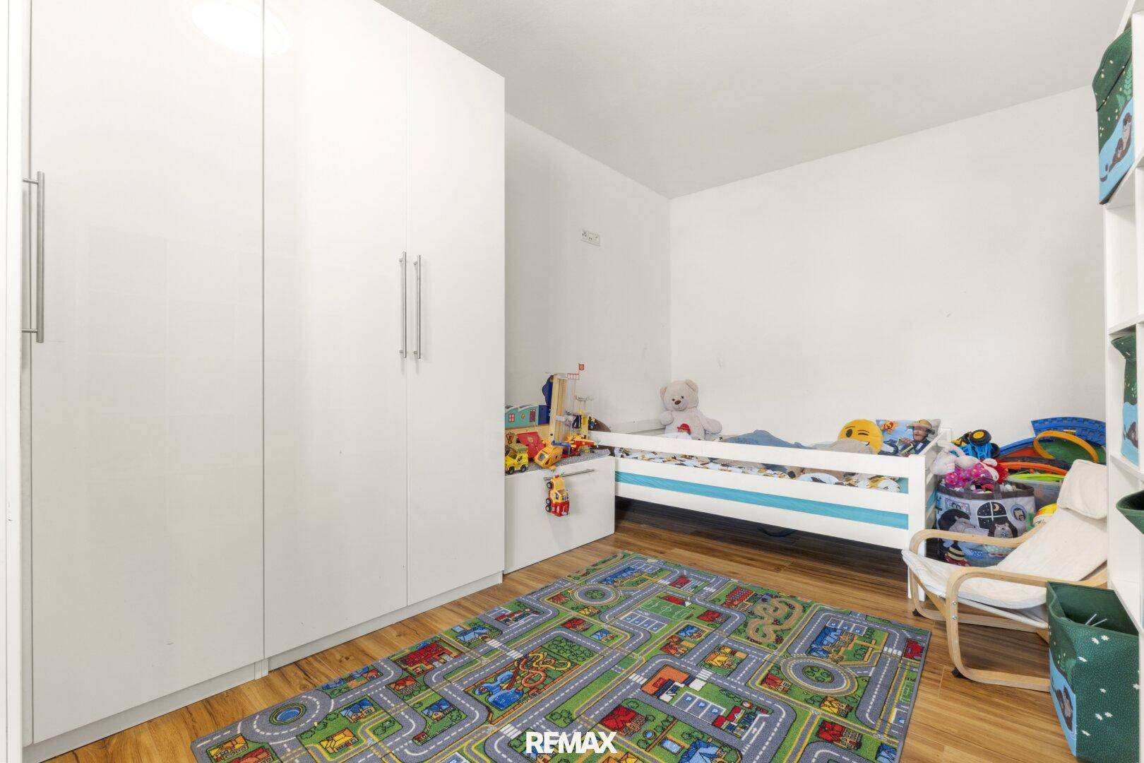 Helles Kinderzimmer mit weißen Schränken, Holzboden und farbenfrohem Spielteppich.