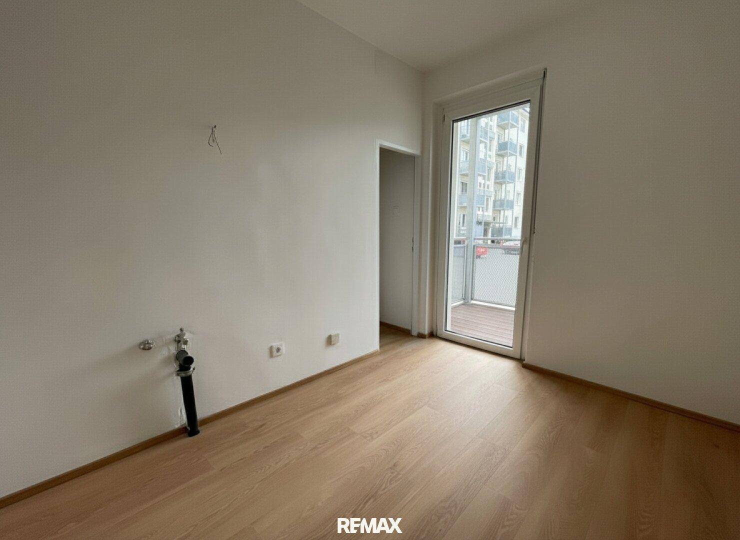 Leerer Raum mit Laminatboden, Balkonzugang und Anschlüssen für eine Küche.