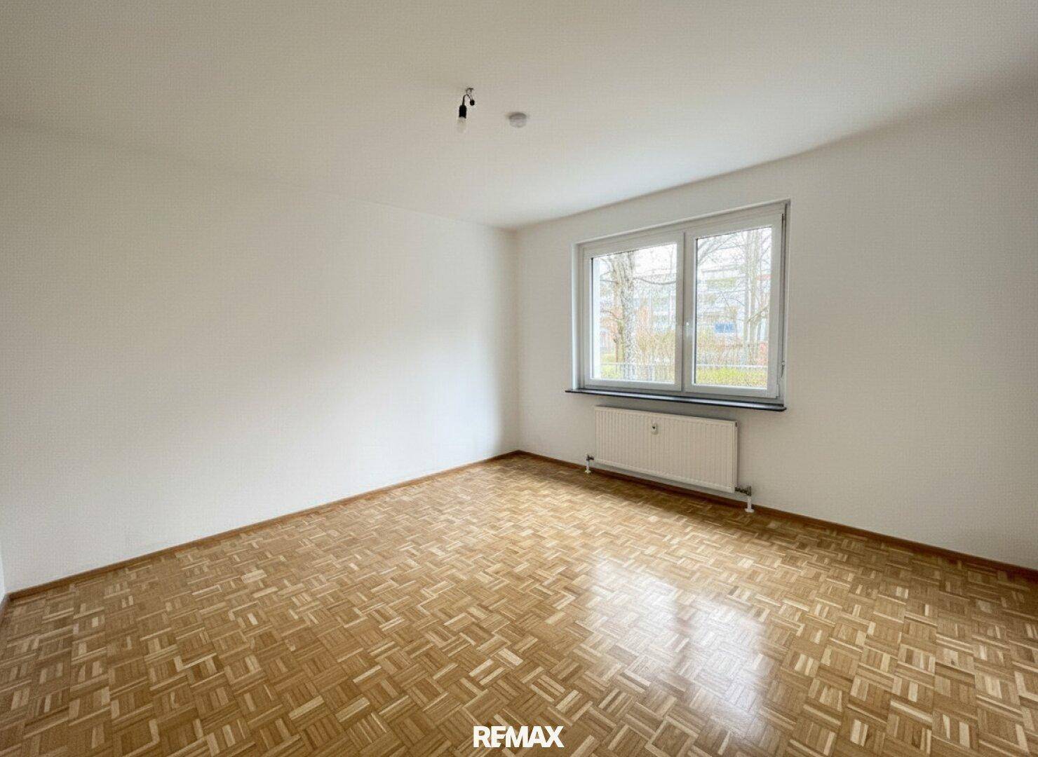 Leerer Raum mit hellem Parkettboden und großem Fenster, das viel Tageslicht hereinlässt.