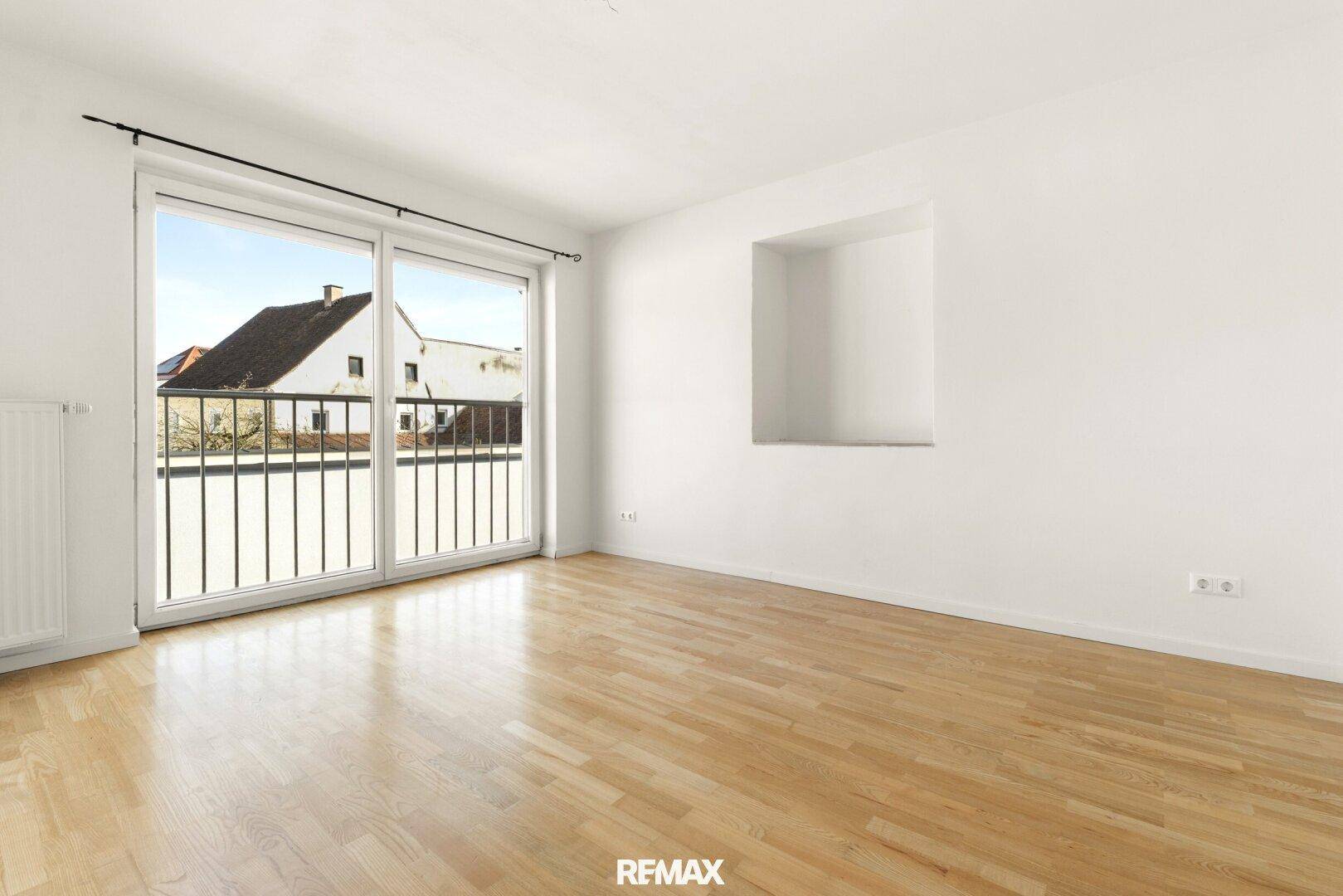 Lichtdurchfluteter Raum mit Holzboden und großer Schiebetür, die zu einem französischen Balkon führt.