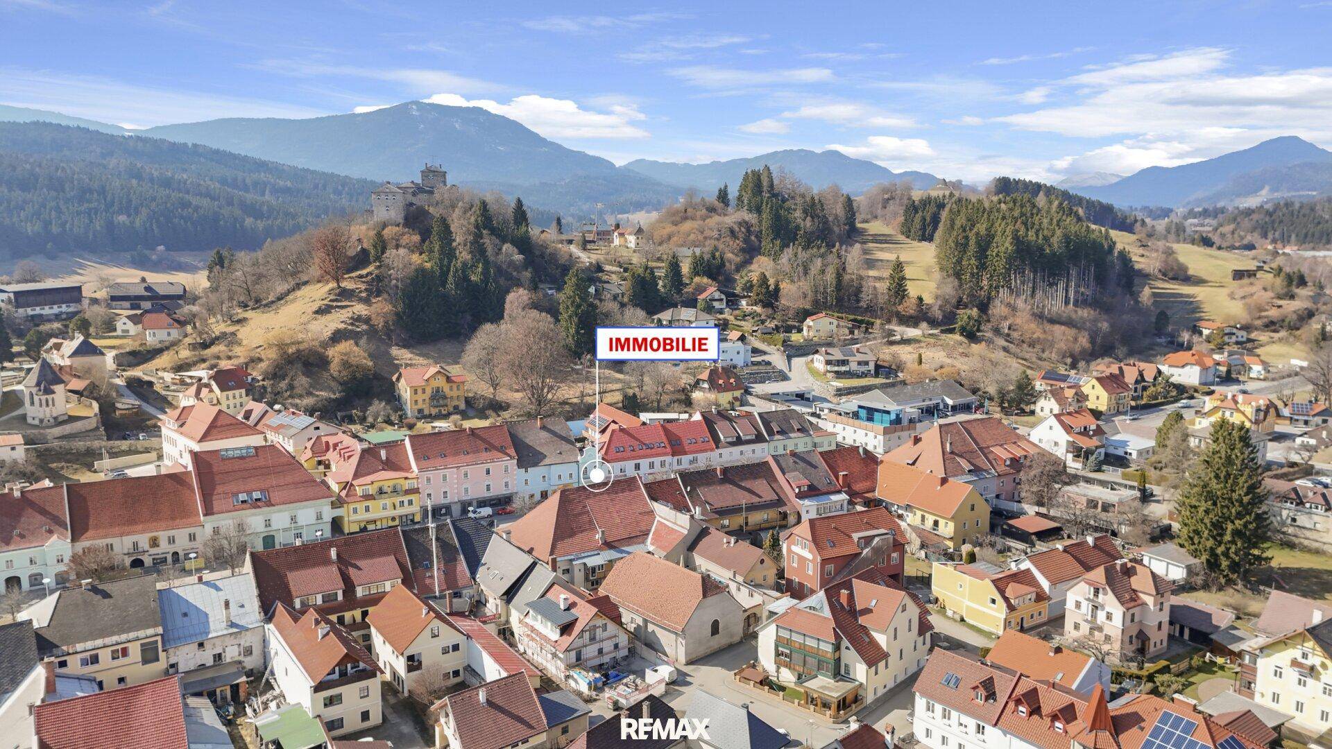Weitläufige Luftaufnahme der Stadt mit historischer Burg auf einem Hügel und Bergkulisse.