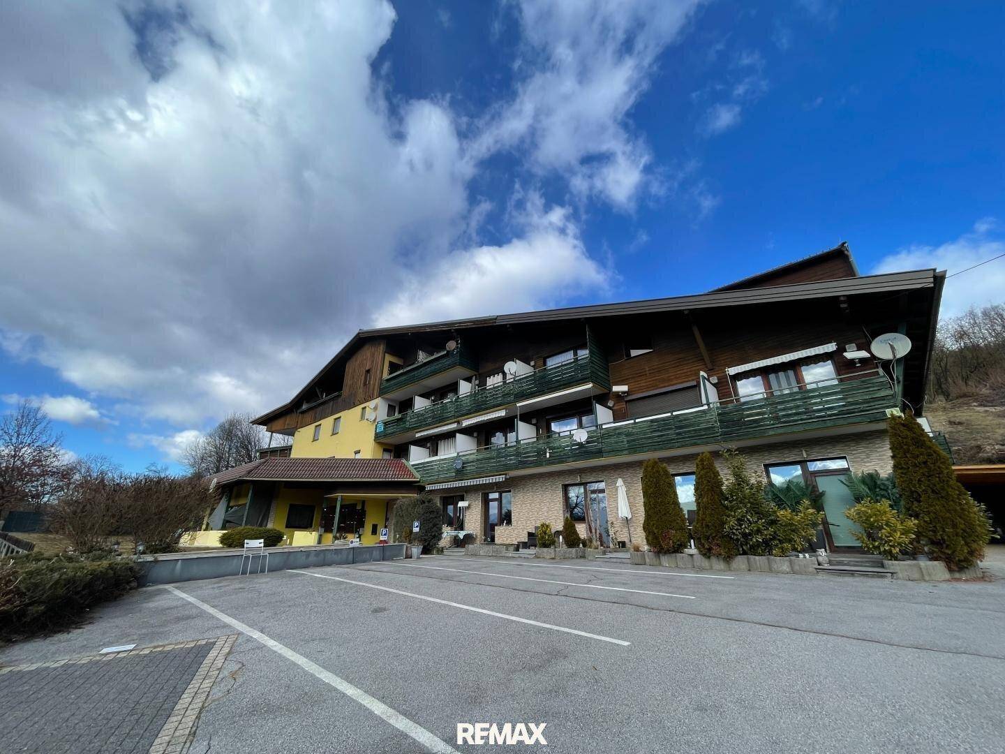 Mehrstöckiges Gebäude mit Balkonen und Parkplätzen im Vordergrund unter bewölktem Himmel.