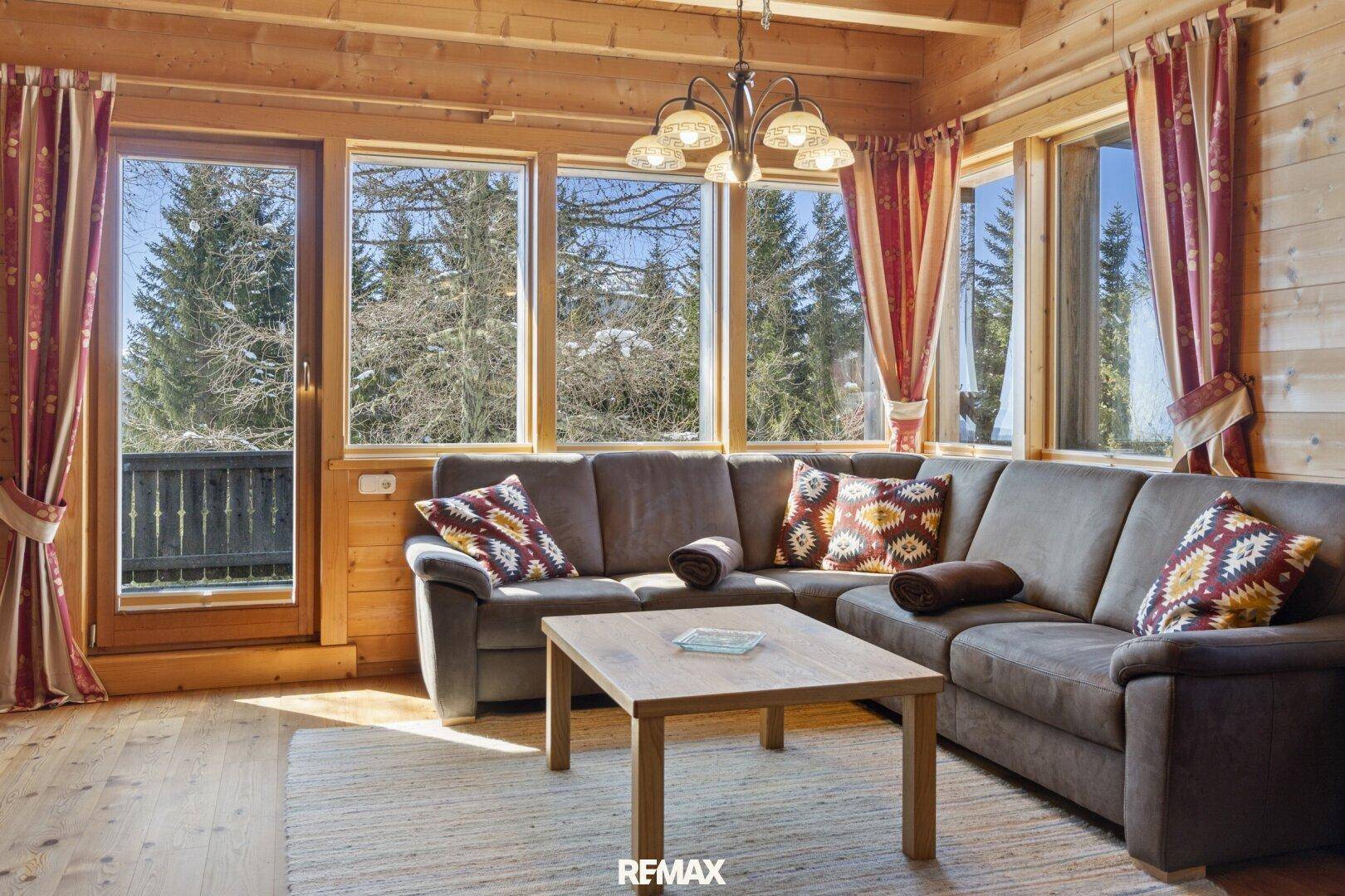 Gemütliches Wohnzimmer im alpinen Stil mit Holzwänden, Ecksofa und großen Fenstern mit Waldblick.