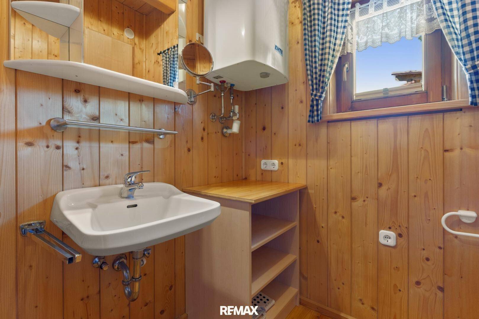 Kleines Badezimmer mit Holzvertäfelung, Waschbecken, Spiegel und einem Fenster mit karierten Vorhängen.