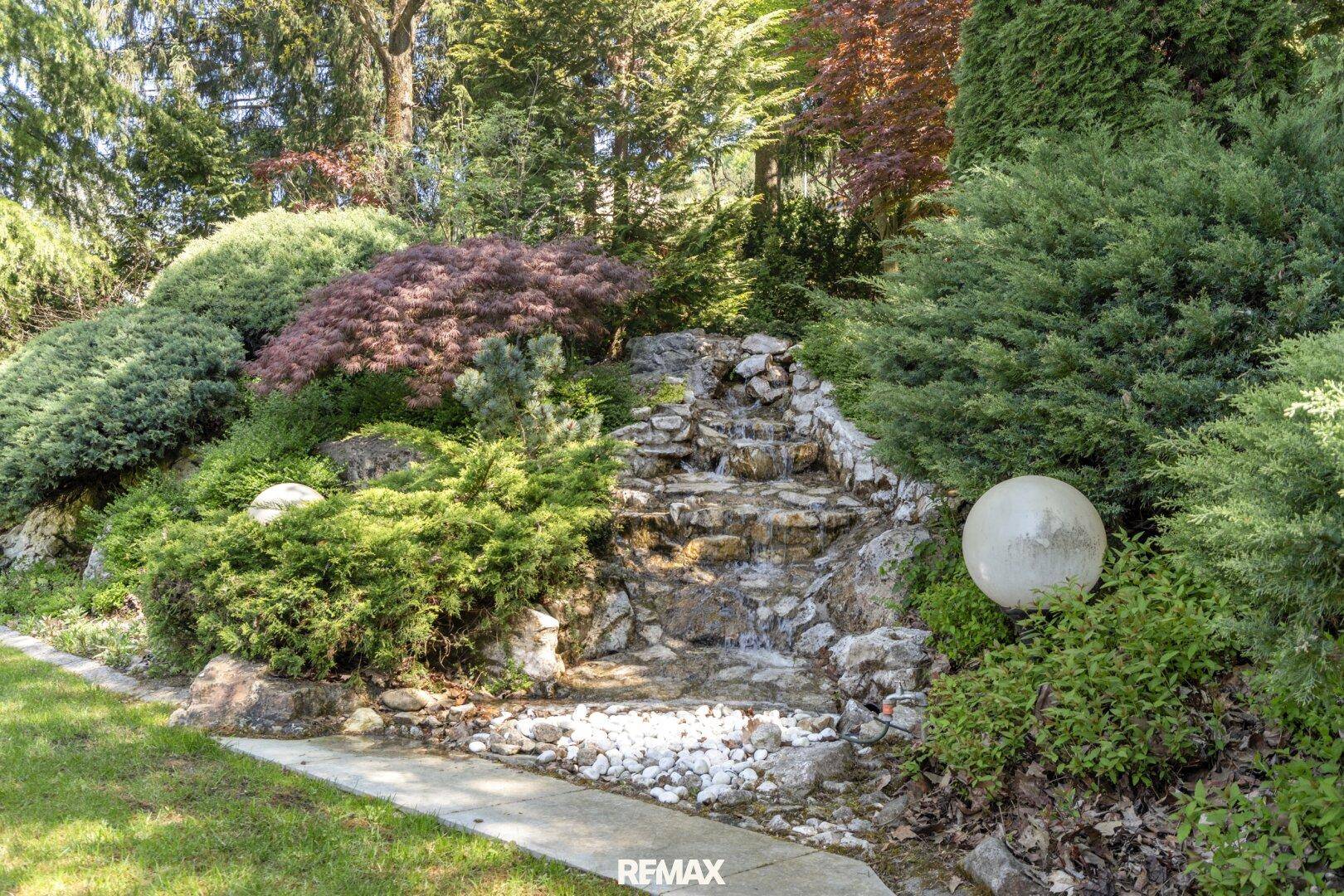 Künstlicher Wasserfall im üppig bepflanzten Gartenbereich mit Steinen und Grünpflanzen.