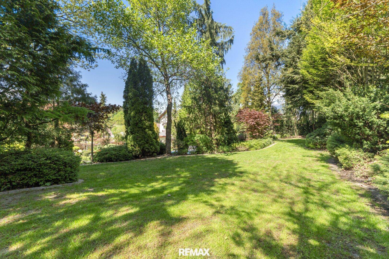 Großer, gepflegter Garten mit weitläufiger Rasenfläche und altem Baumbestand unter blauem Himmel.