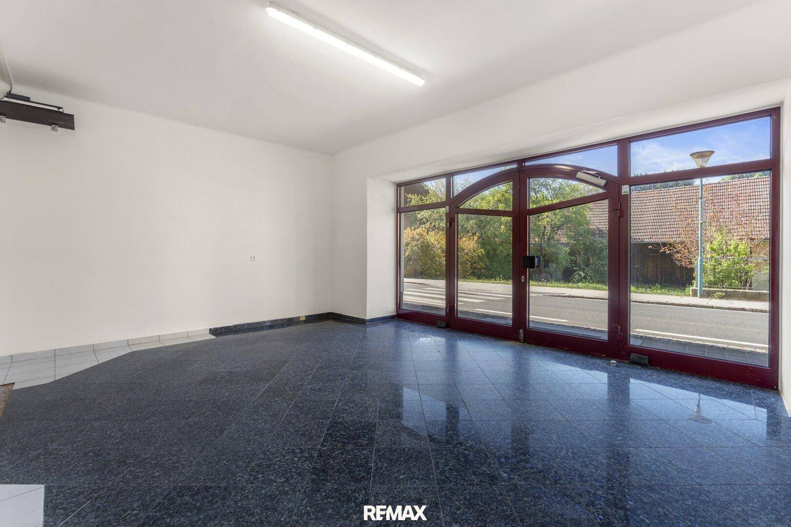 Heller Raum mit großen Glasfronten zur Straße und dunklem Fliesenboden, ideal für Büro oder Laden.
