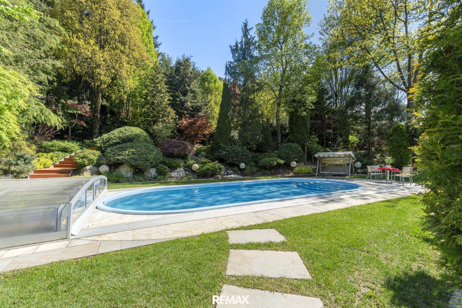Großer ovaler Swimmingpool im Gartenbereich, umgeben von Rasen und Bäumen.