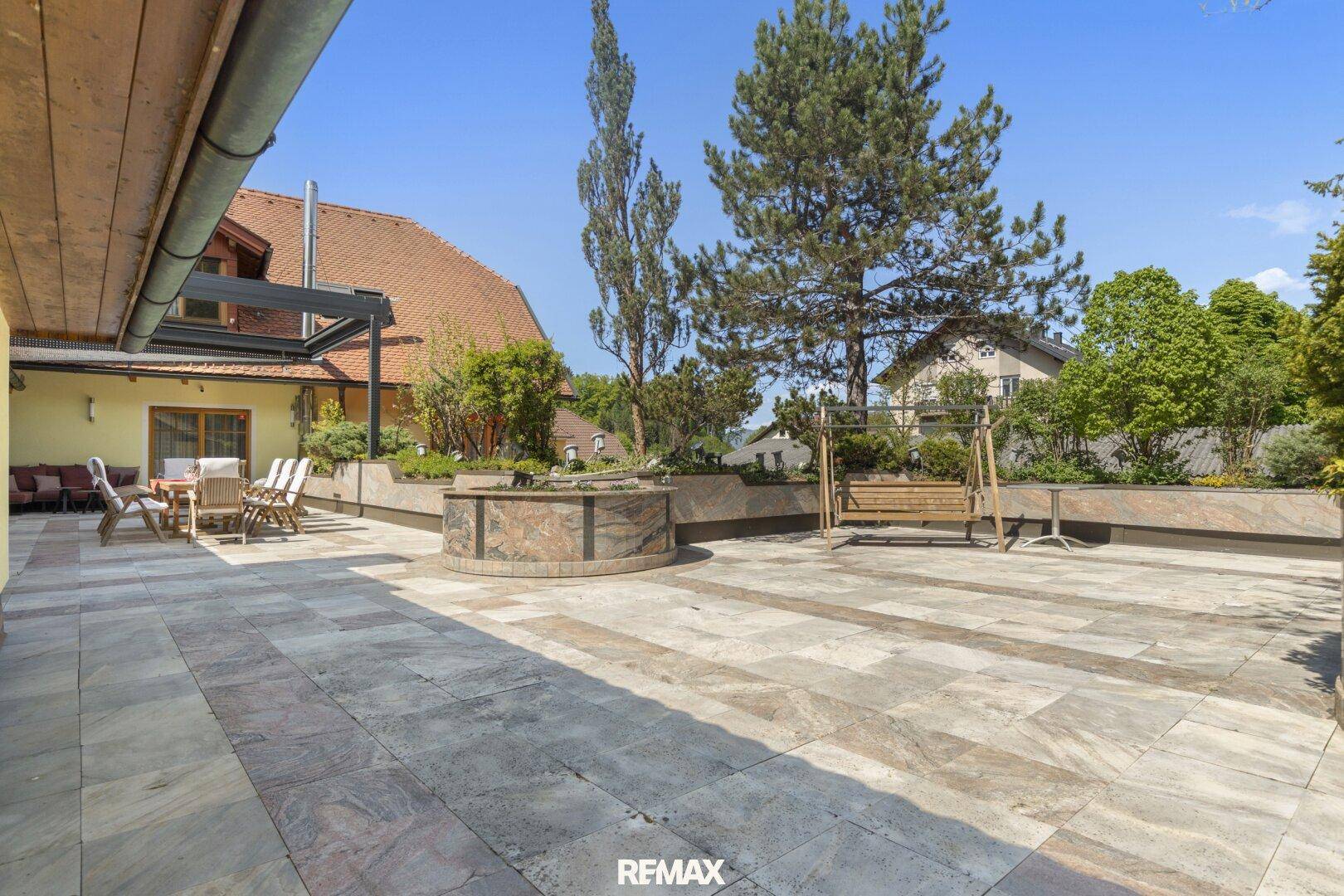 Geräumige geflieste Terrasse mit Sitzgelegenheiten, Schaukel und Pflanzenbeeten unter blauem Himmel.