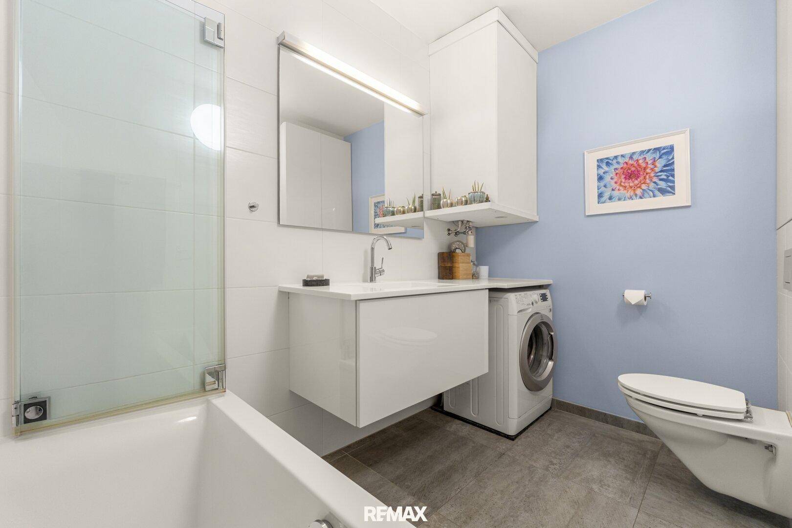 Modernes Badezimmer mit Waschbecken, Toilette, Waschmaschine und einer Badewanne mit Duschabtrennung.