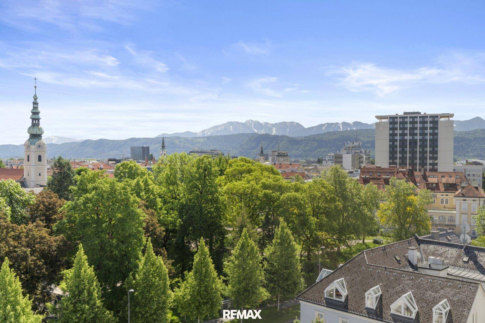 Panoramablick über die Stadt mit grünen Bäumen und majestätischen Bergen im Hintergrund unter blauem Himmel.