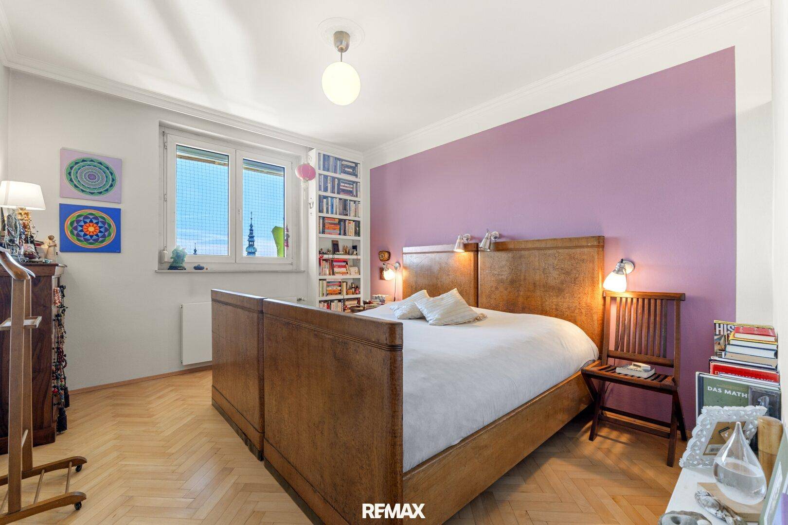 Gemütliches Schlafzimmer mit großem Holzbett, violetter Akzentwand und einem Fenster, das Tageslicht hereinlässt.