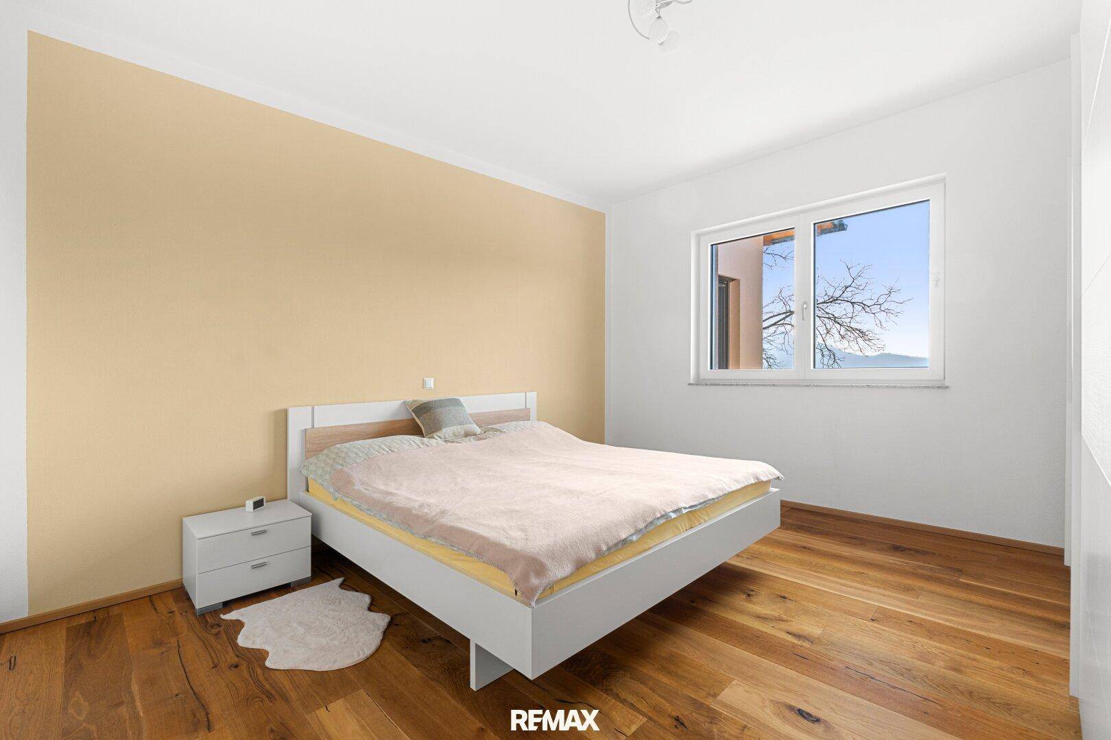 Gemütliches Schlafzimmer mit Holzboden, beiger Akzentwand und Fenster mit Blick ins Grüne.