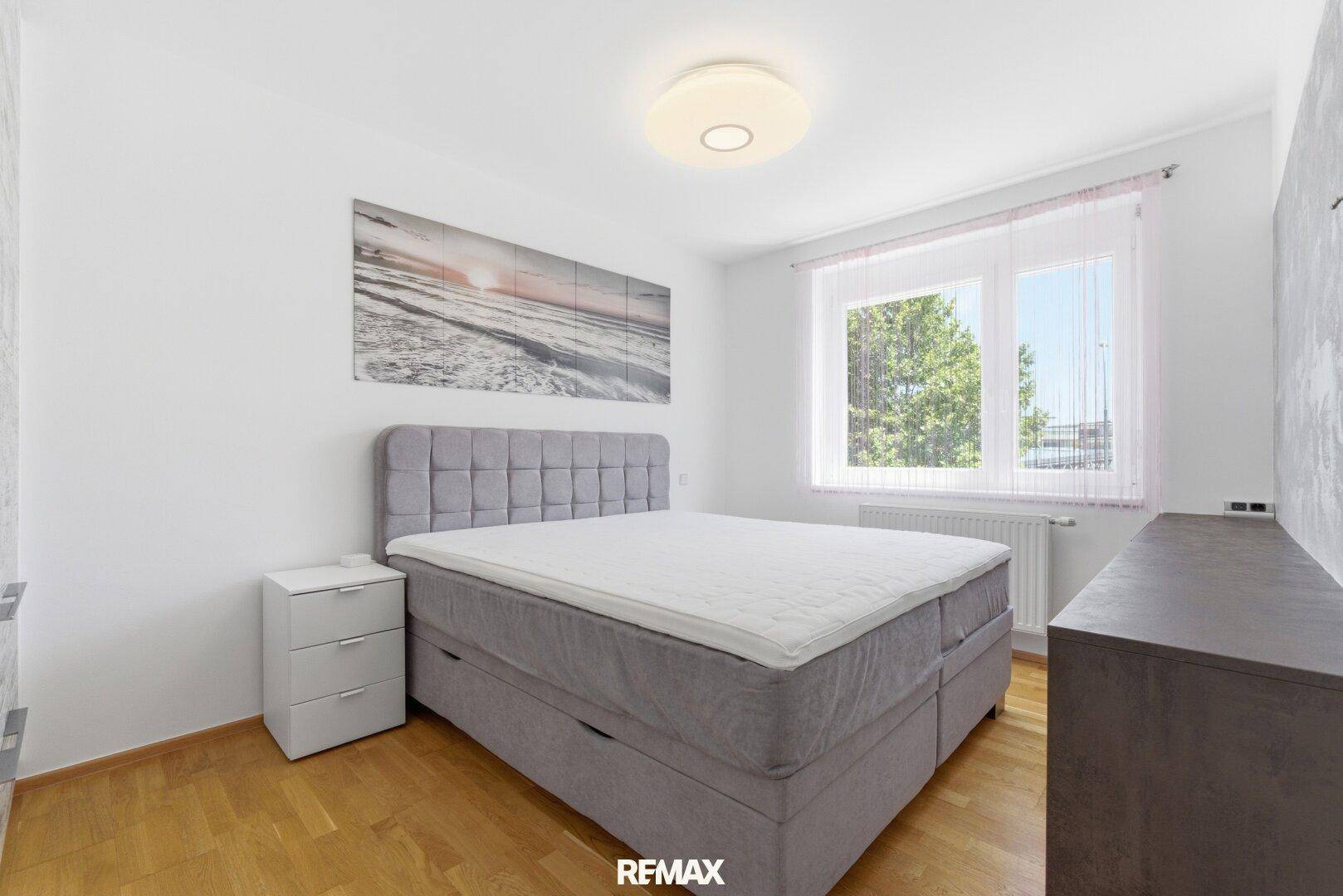 Helles Schlafzimmer mit großem Boxspringbett, Holzboden und Fenster mit Blick ins Grüne.