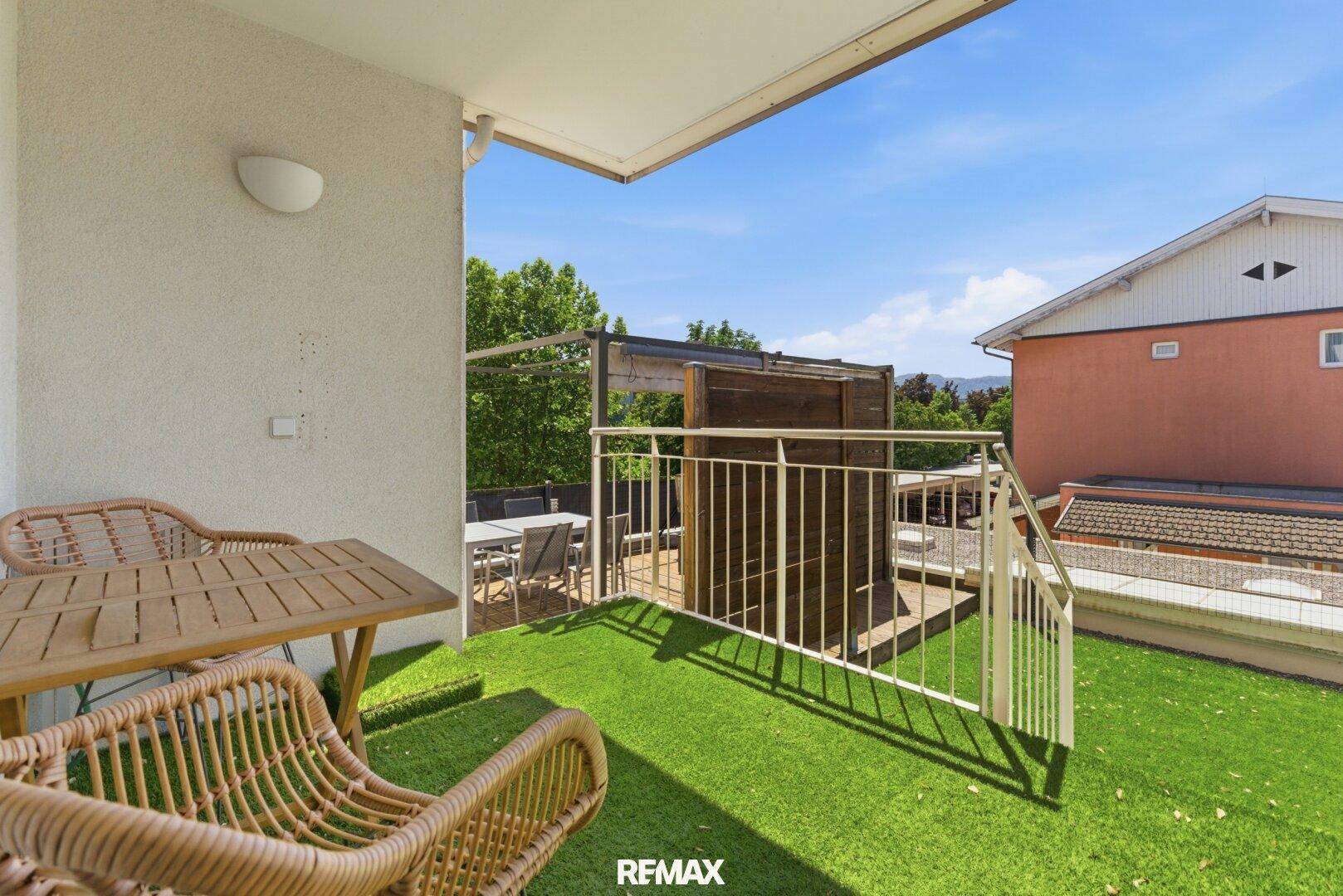 Sonniger Balkon mit Kunstrasen, Sitzgelegenheiten und Blick auf die umliegenden Häuser.