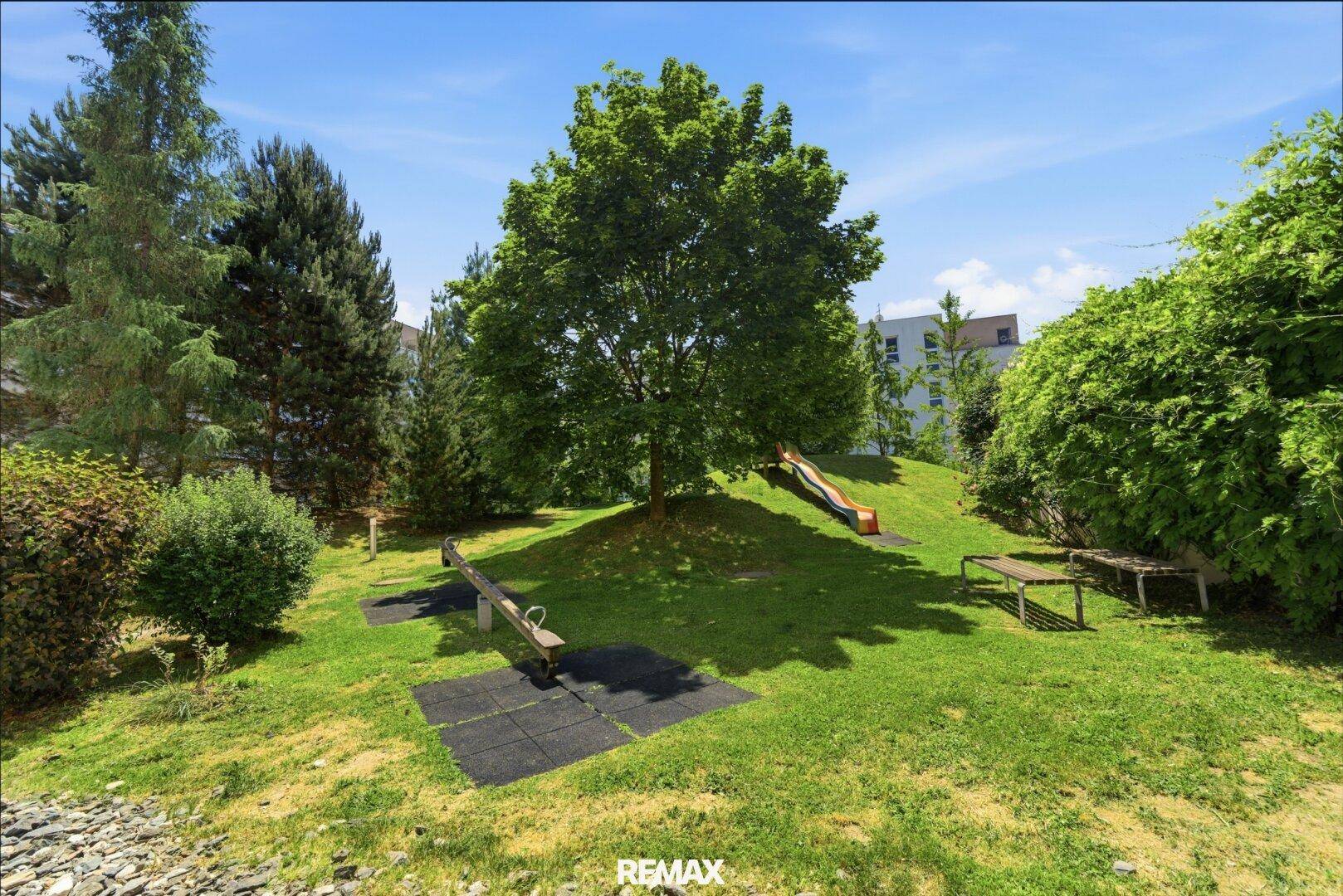 Grüner Spielplatz mit Rutsche und Wippe, umgeben von Bäumen und Sträuchern.