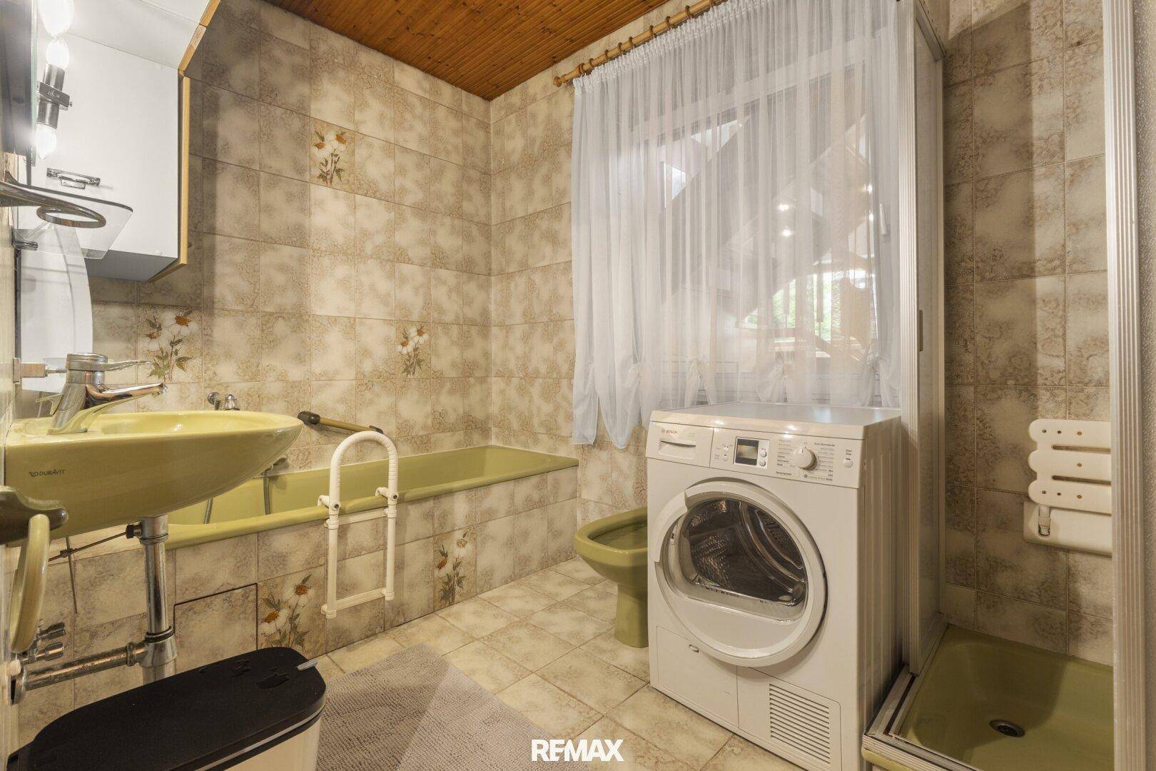 Badezimmer mit Badewanne, Dusche, Waschmaschine und Fliesen im Retro-Stil, mit Haltegriffen.