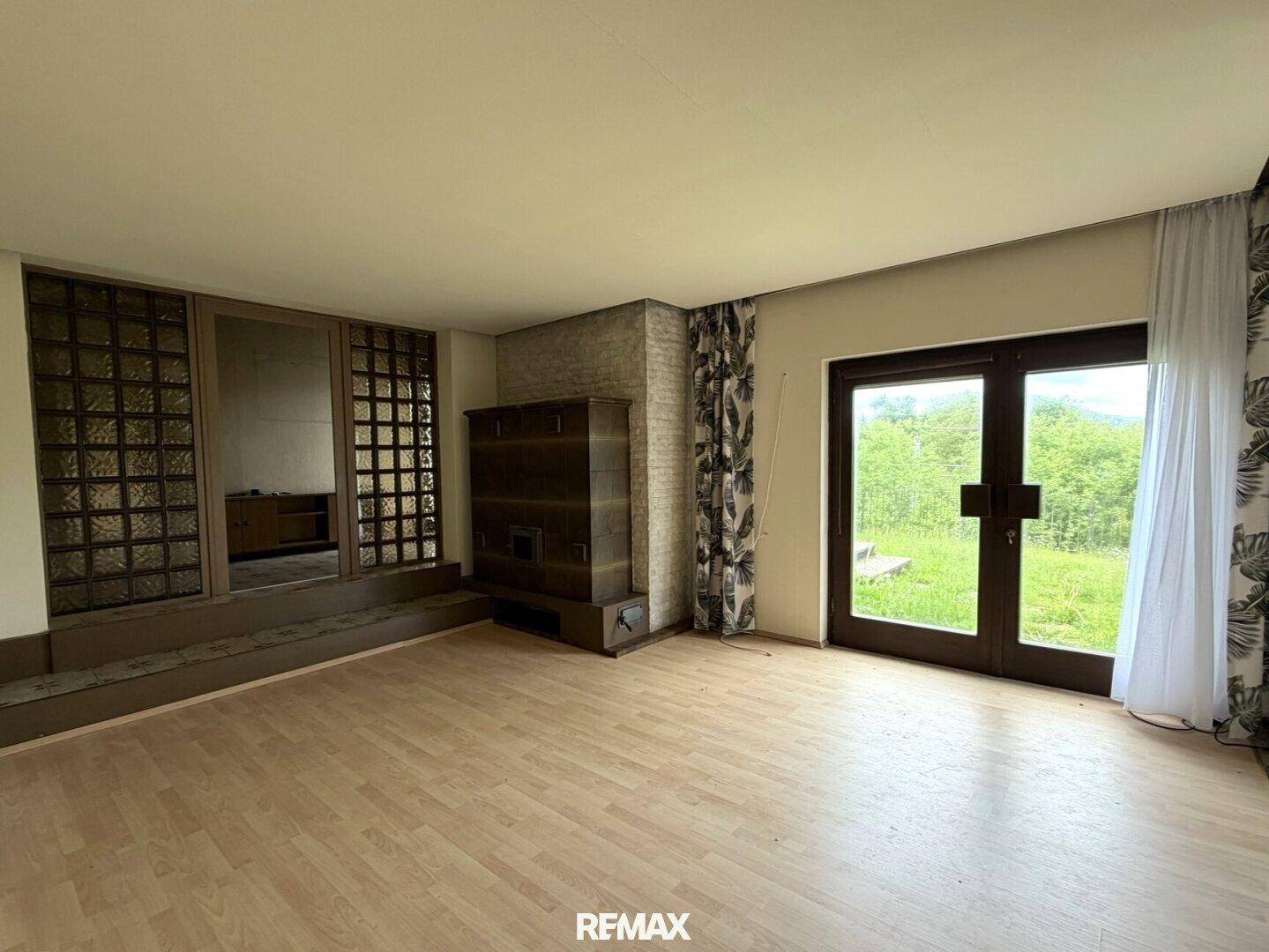 Geräumiges Wohnzimmer mit Holzboden, Kachelofen und Zugang zum Garten durch große Türen.