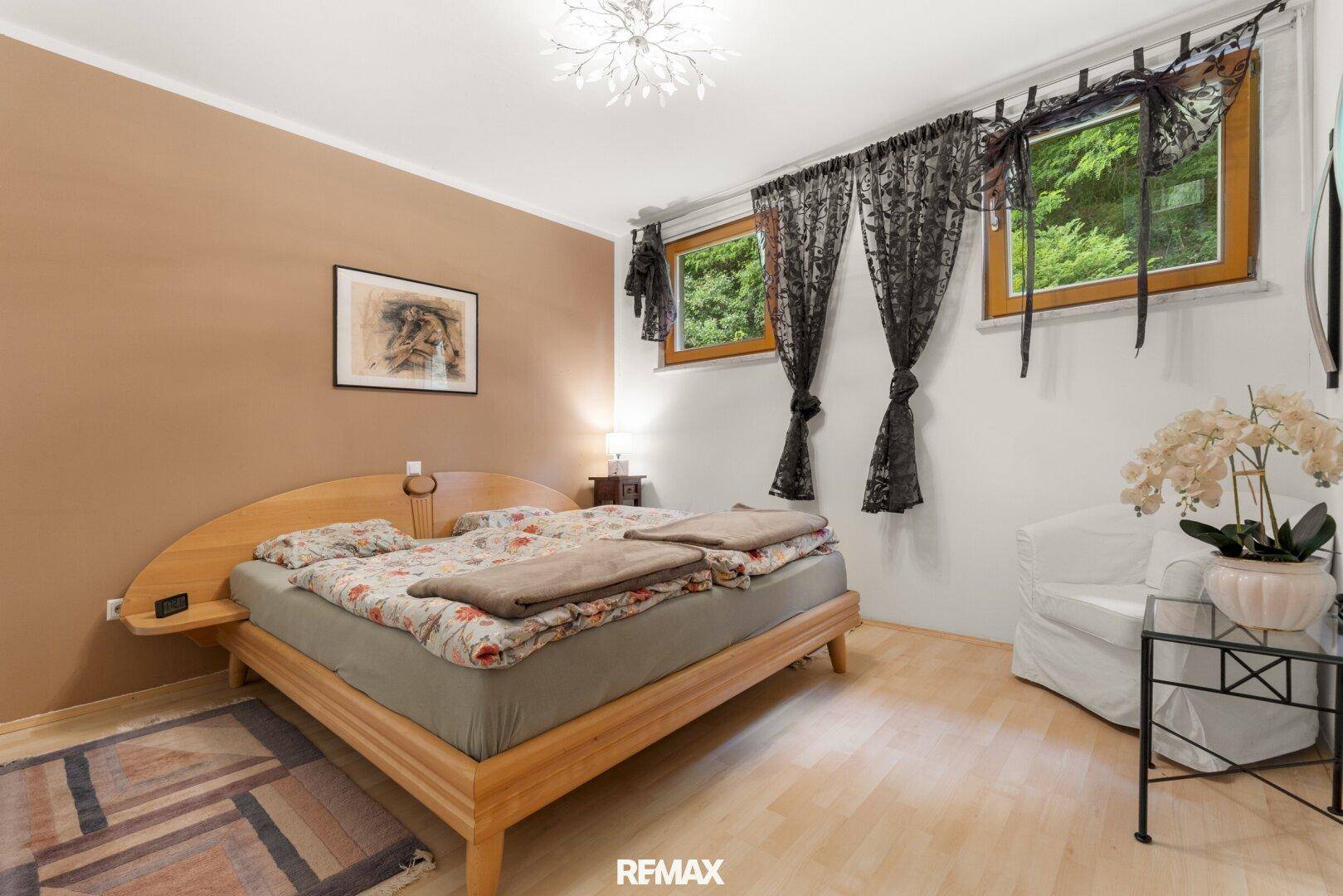 Helles Schlafzimmer mit Holzbett, Parkettboden und Blick ins Grüne durch die Fenster.