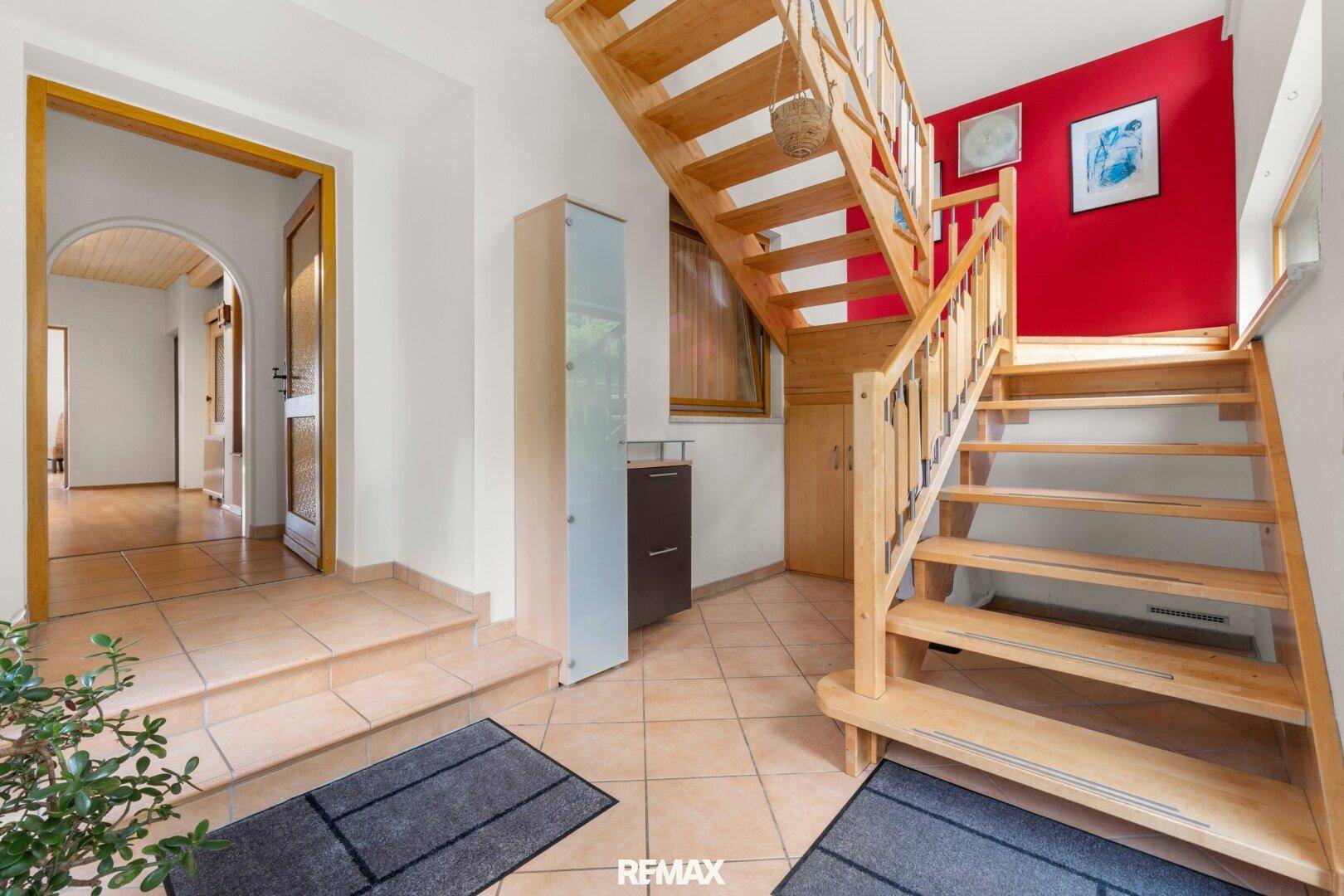 Heller Eingangsbereich mit Holztreppe, Terrakotta-Fliesenboden und roter Akzentwand.