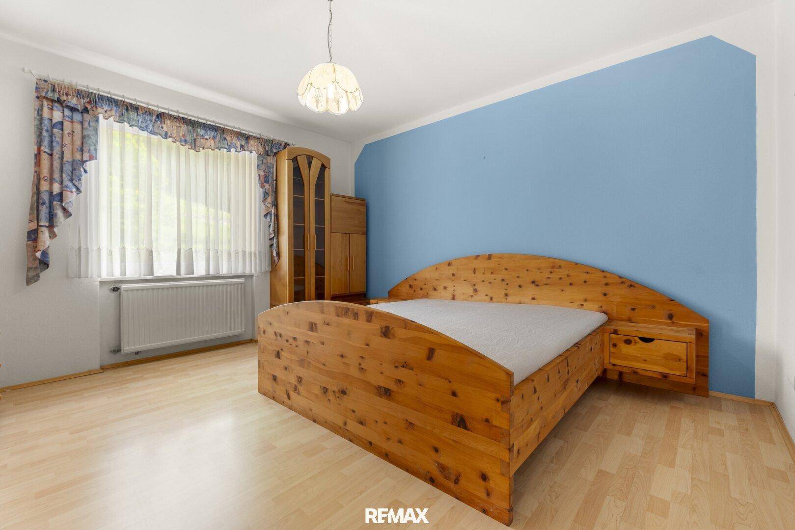 Schlafzimmer mit hellem Holzbett, Kleiderschrank und einer blauen Akzentwand, gut beleuchtet.