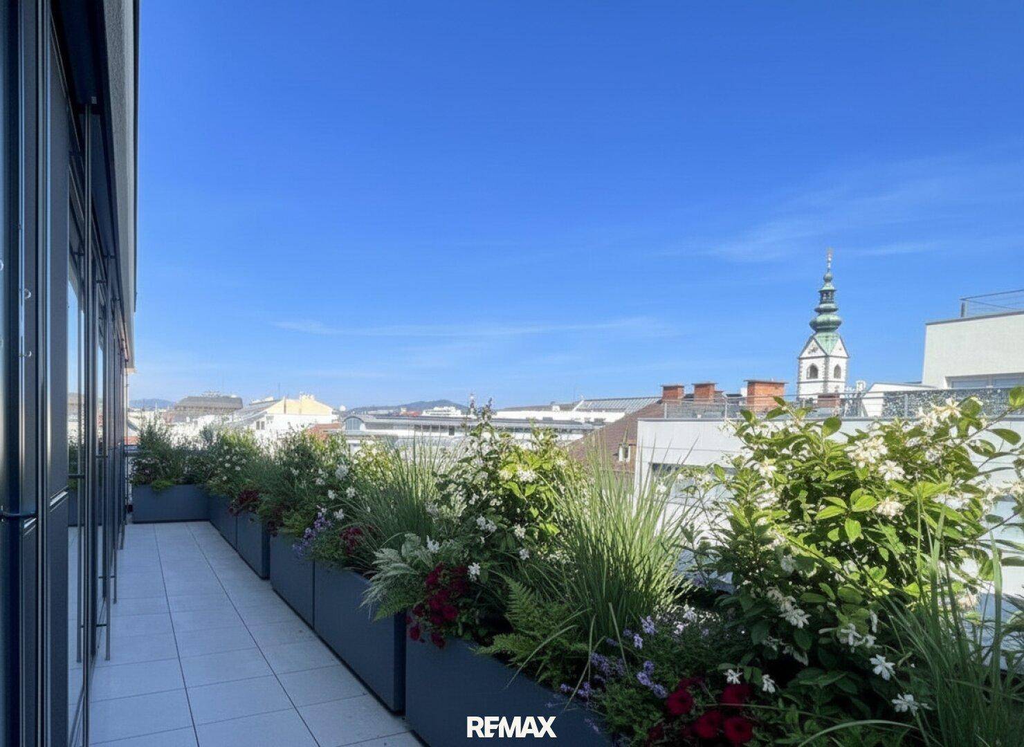 Begrünte Dachterrasse mit Blumenkästen und Blick auf die Dächer der Stadt und einen Kirchturm.