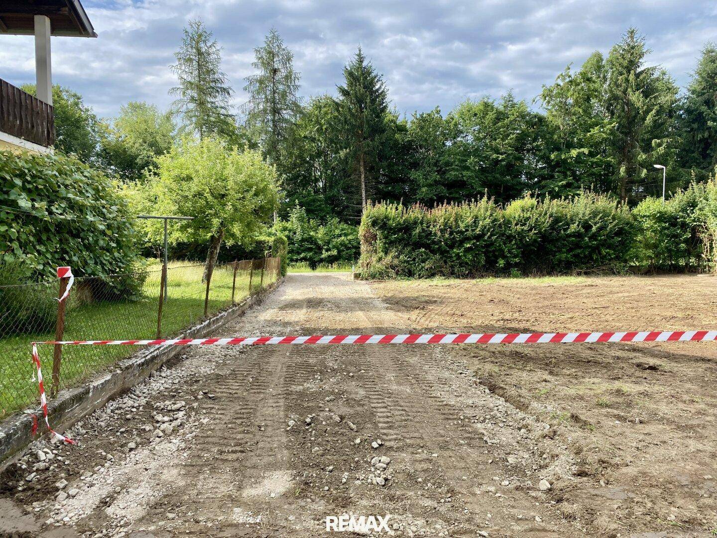 Zufahrtsweg aus Schotter zu einem Grundstück, gesichert mit rot-weißem Absperrband und Bäumen.