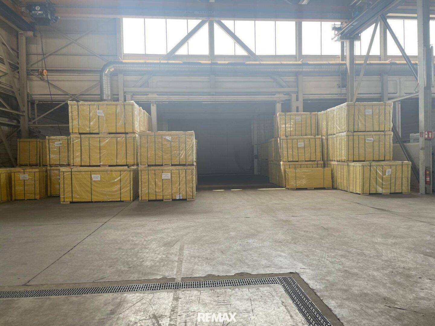 Große Lagerhalle mit gestapelten Paletten und einem breiten Zugangstor im Hintergrund.