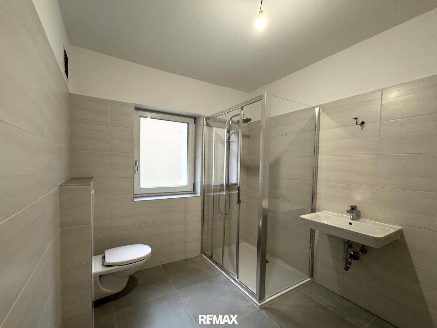 Modernes Badezimmer mit hellgrauen Fliesen, Duschkabine, WC und Fenster für Tageslicht.